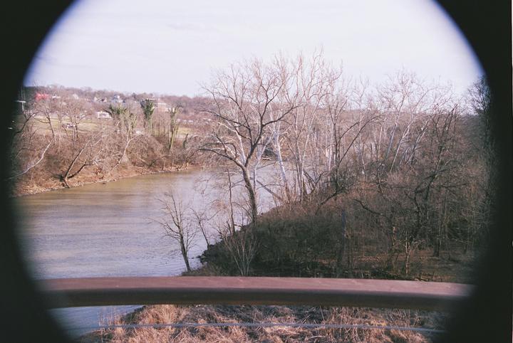A river flows past a wooded bank with bare trees and a distant town under a bright sky, viewed through a circular frame.