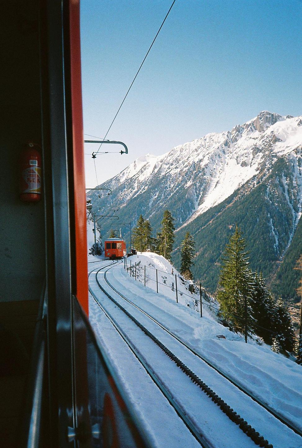 An orange train travels on a snowy mountain track with pine trees and a snow-covered mountain in the background.
