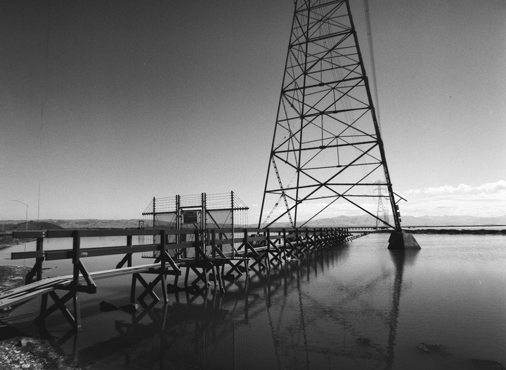 A large metal tower stands in water next to a wooden fence and a barbed wire fence.