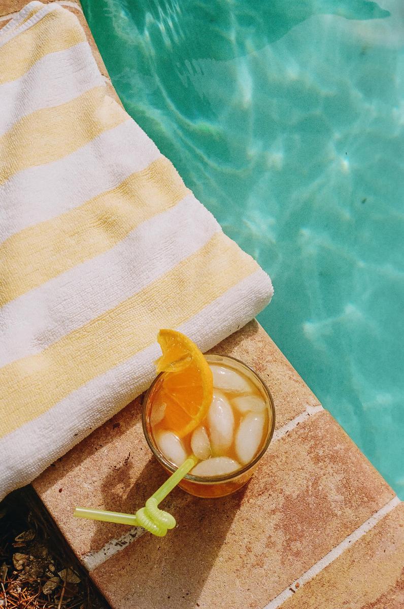 A glass of iced tea with an orange slice and a green straw sits next to a striped towel by a swimming pool.