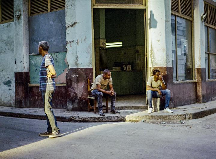 Three men are on a street corner with two sitting in front of a building and one standing on the street.