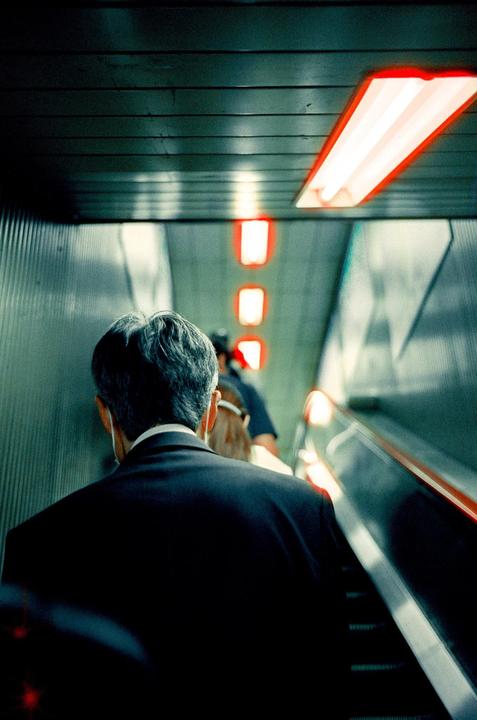 A man in a suit ascends an escalator with red lights on the ceiling.