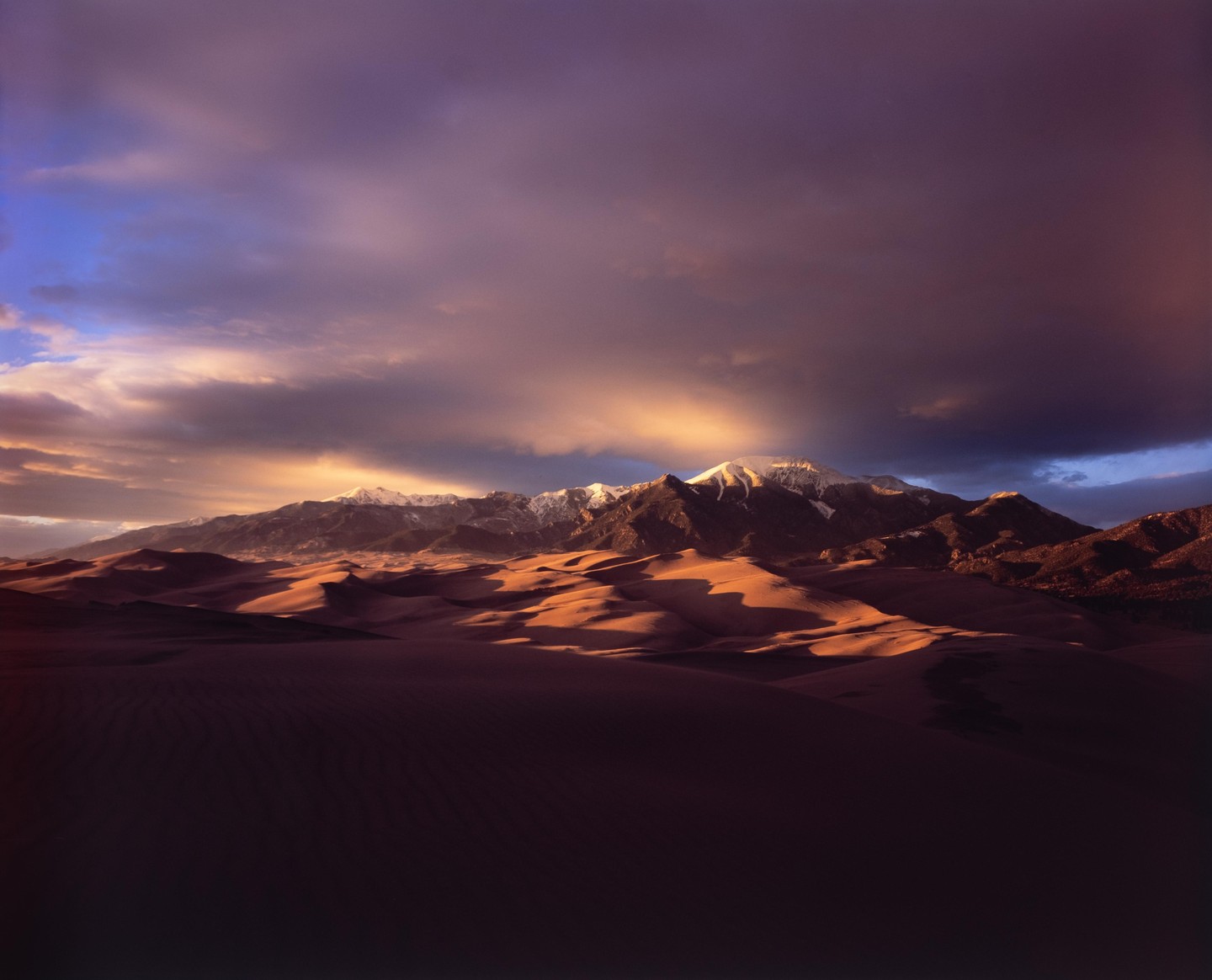 A dramatic sunset illuminates sand dunes and snow-capped mountains under a cloudy sky.