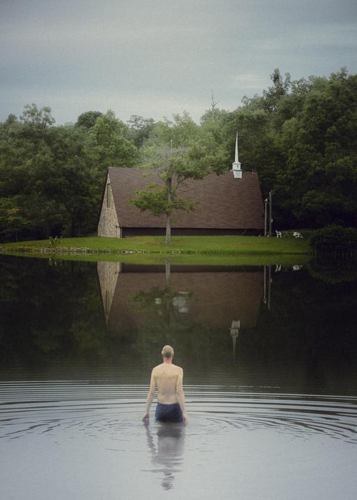 A man stands in a body of water with a church and trees reflected in the water.