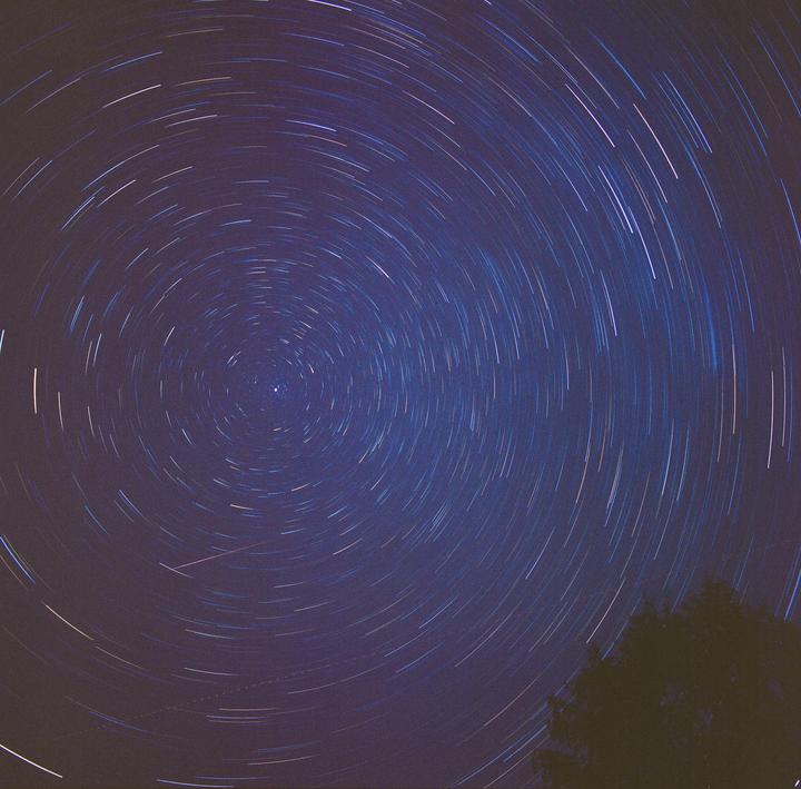 Star trails swirl around a central point in the night sky, with the silhouette of a tree visible in the bottom right corner.