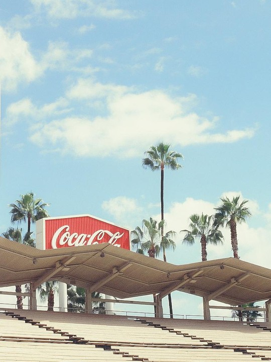 A red Coca-Cola sign is visible above stadium seating and palm trees under a blue sky with clouds.