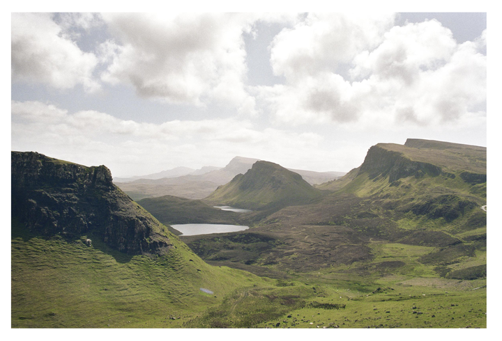 A landscape view shows green, rocky mountains with small lakes under a cloudy sky.