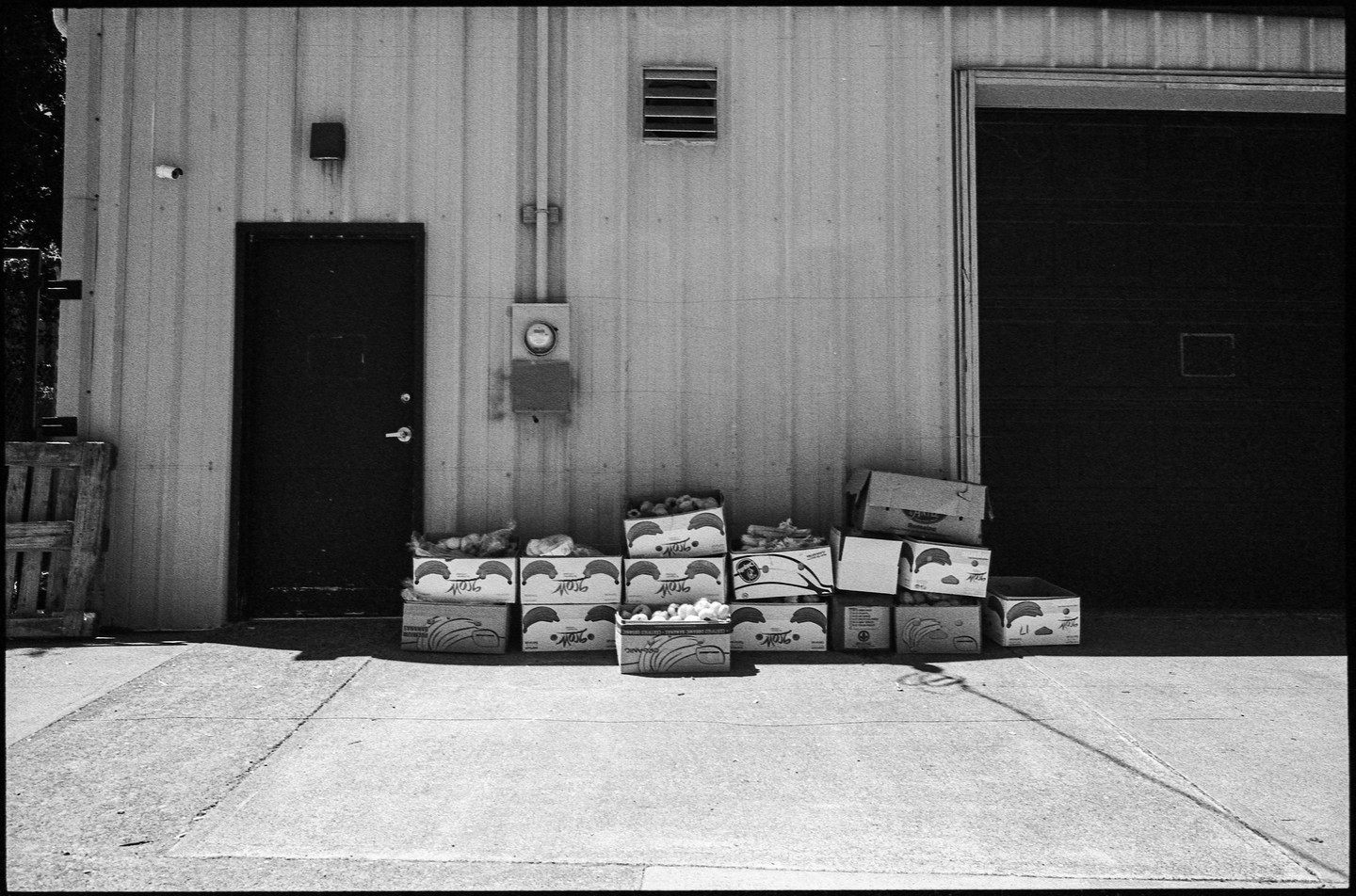 A black and white photograph shows a building with a door, a garage door, and boxes of produce stacked outside.