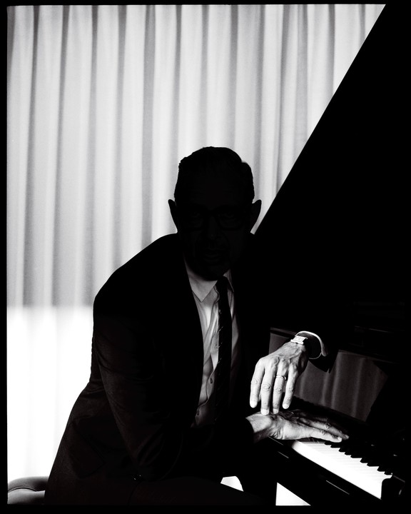 A man in a suit sits at a piano with his hands on the keys, with a curtain in the background.