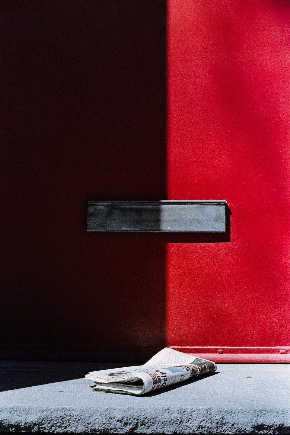 A newspaper rests on a concrete step in front of a red door with a mail slot, partially illuminated by a strong shadow.