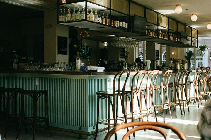A long bar with many stools is lit by sunlight streaming in from large windows.