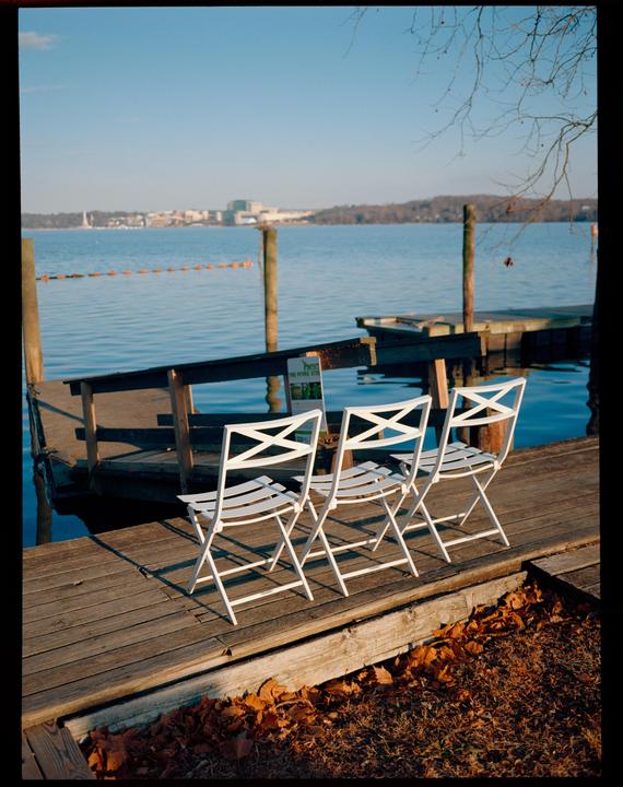 Three white folding chairs sit on a wooden dock next to a body of water with a distant shoreline.