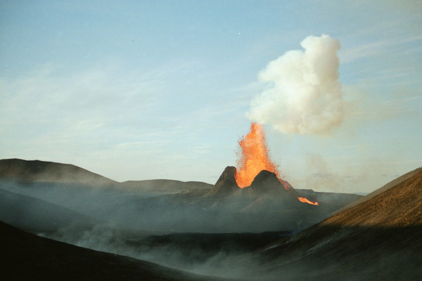 A volcano erupts, spewing lava and smoke into the sky, surrounded by hills and a hazy valley.
