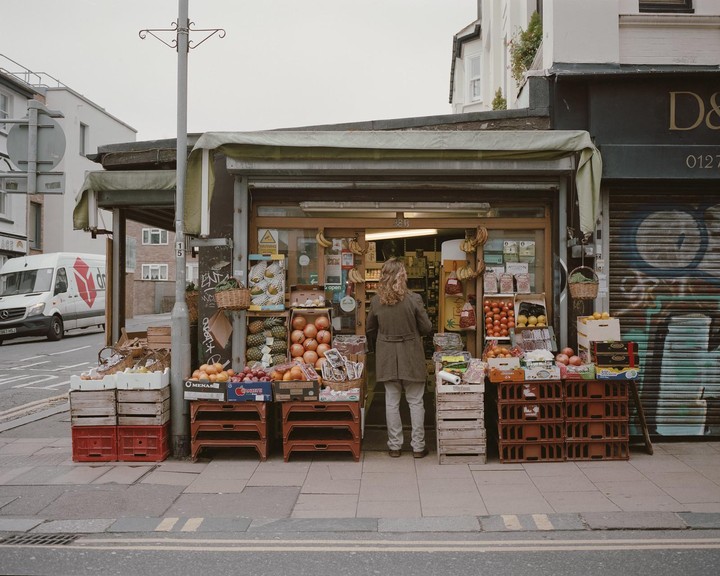A woman stands in front of a fruit and vegetable stand on a city street.
