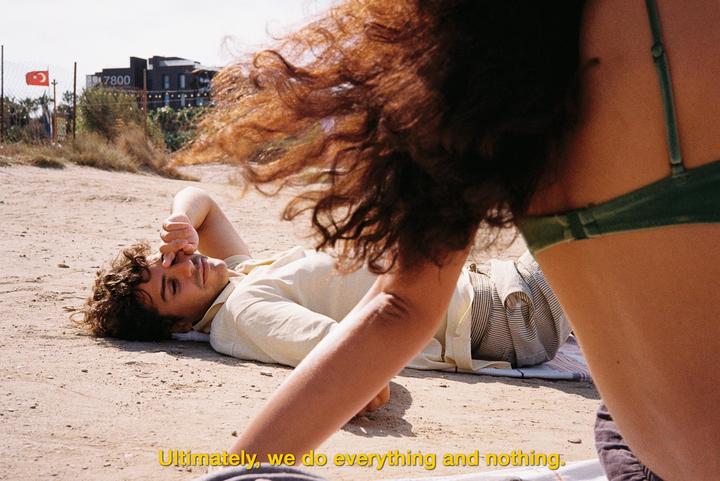 A man lies on a sandy beach with a woman's back and hair in the foreground.