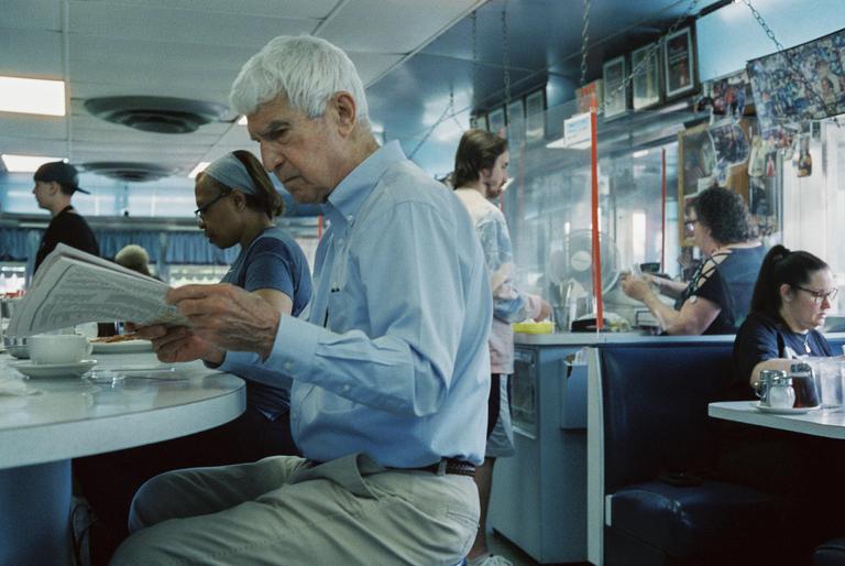 A man with white hair reads a newspaper at a diner counter while other patrons sit and stand in the background.