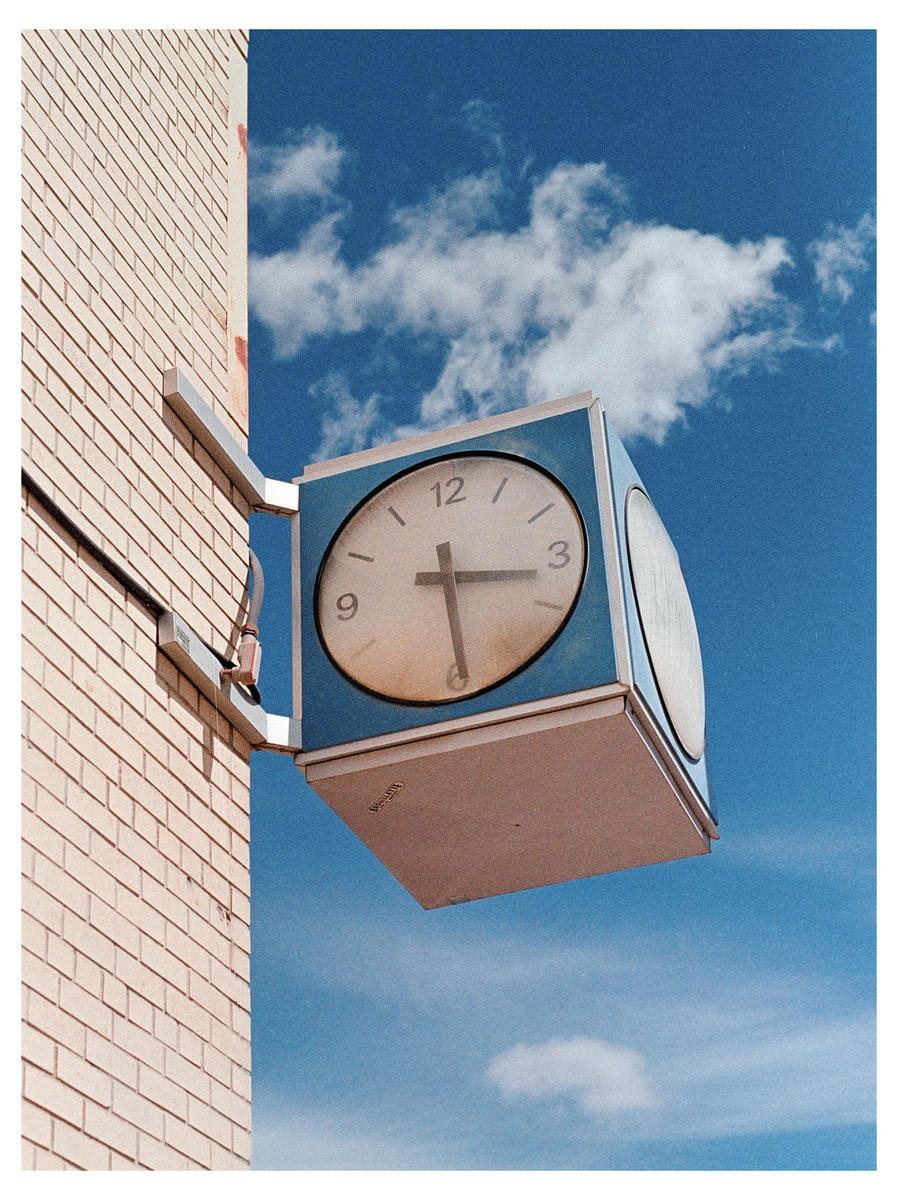 A square clock with a round face is mounted on a brick building against a blue sky with white clouds.