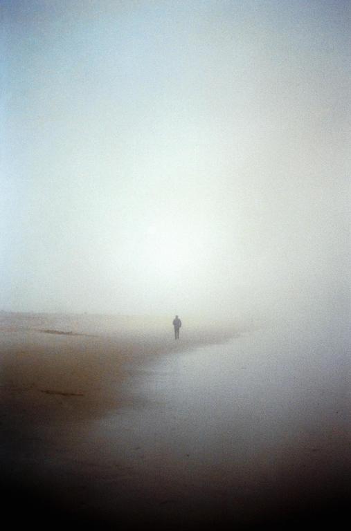 A lone man walks along a foggy beach.
