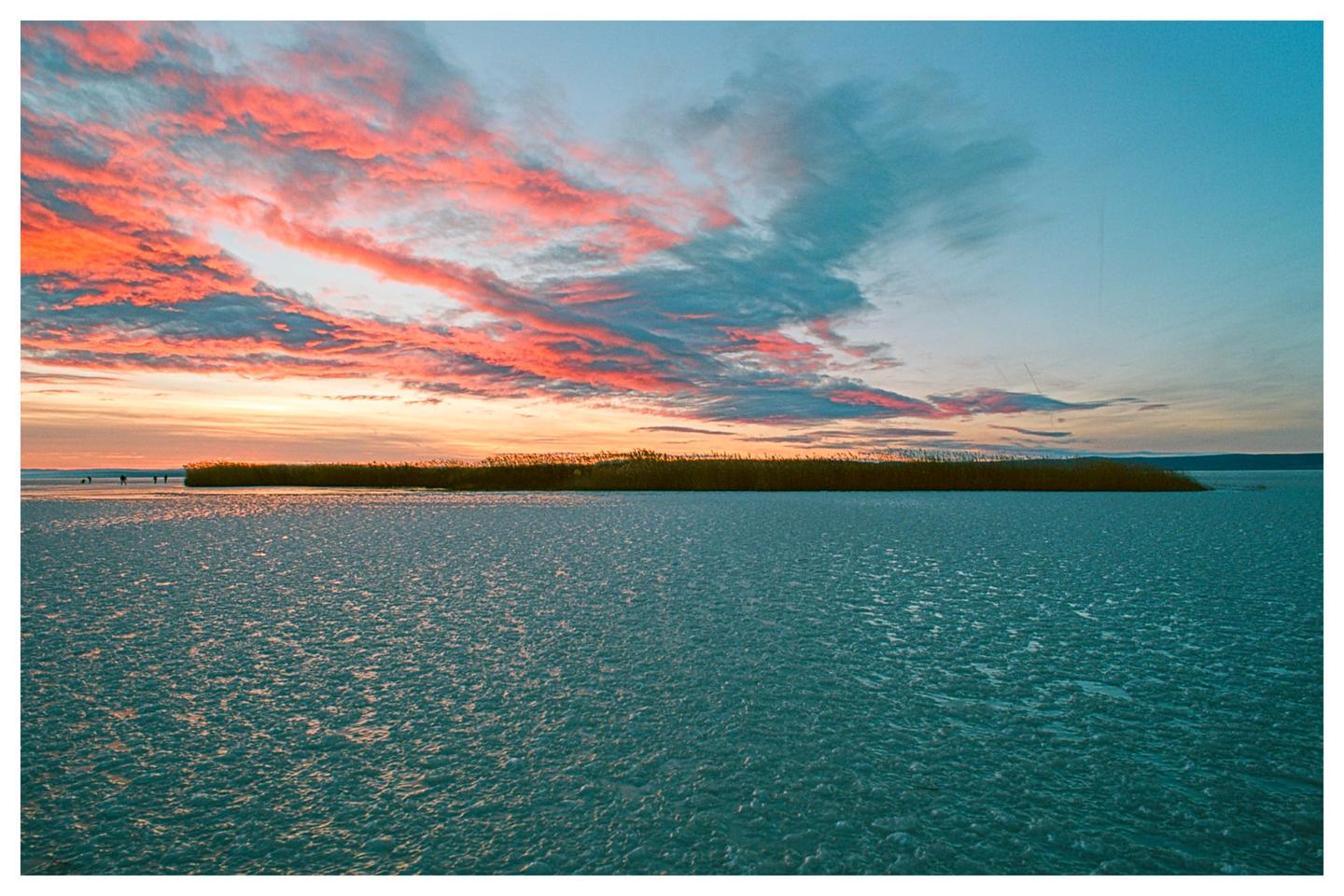 A frozen lake with a strip of reeds in the distance is illuminated by the vibrant colors of a sunset sky.