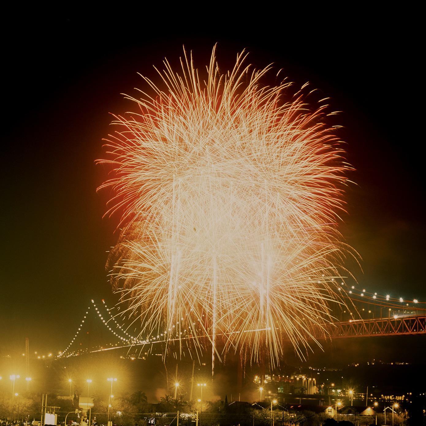Fireworks explode over a city with a suspension bridge at night.