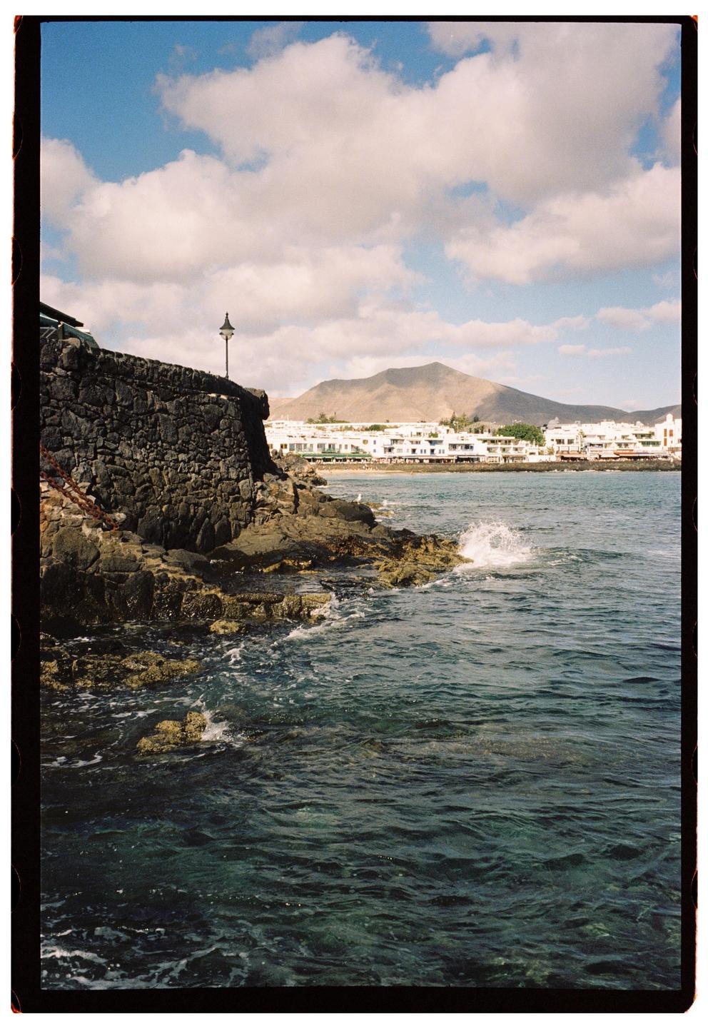 A rocky coastline with a town and mountain in the background under a cloudy sky.