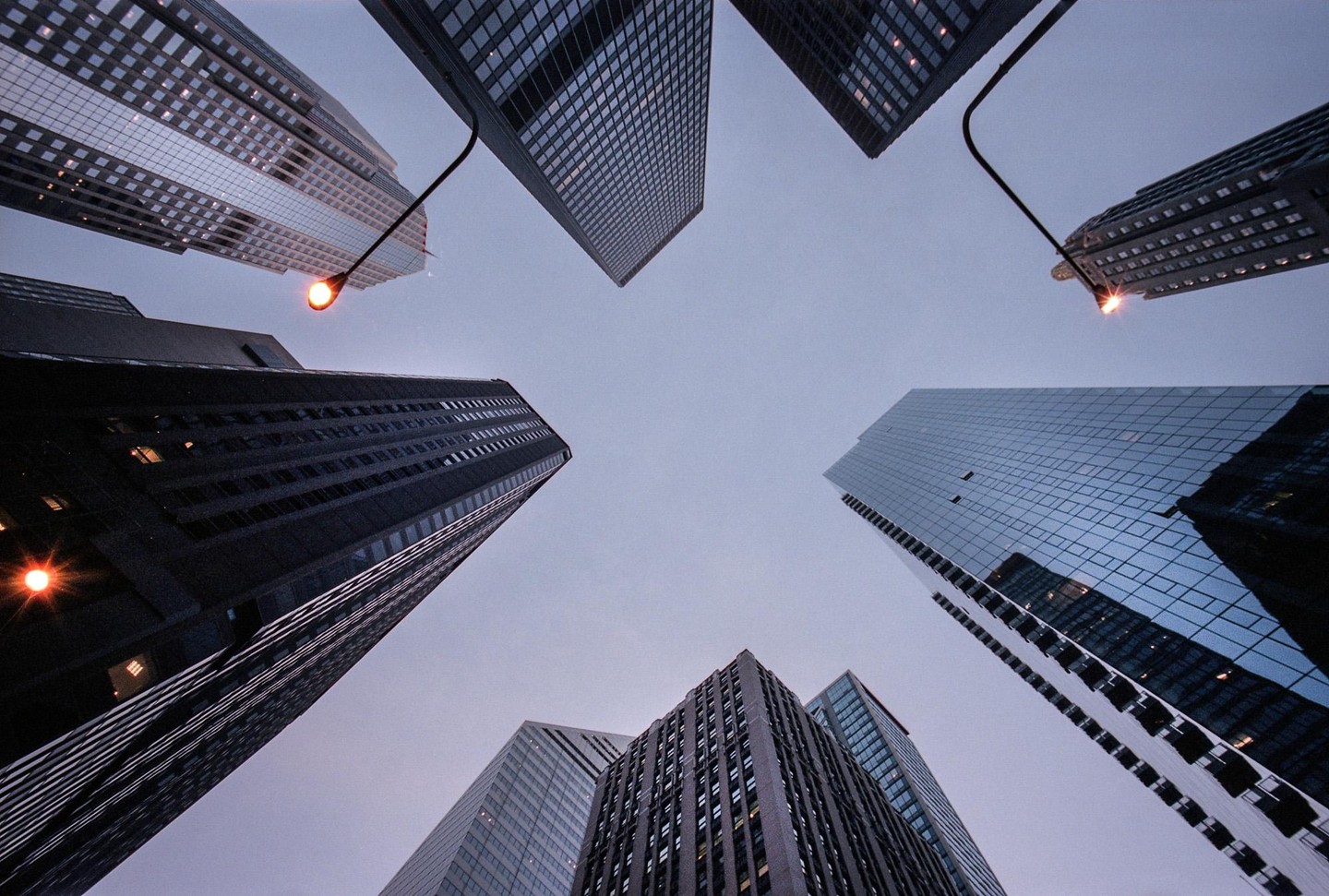 Looking up at several tall buildings and two streetlights against a pale sky.