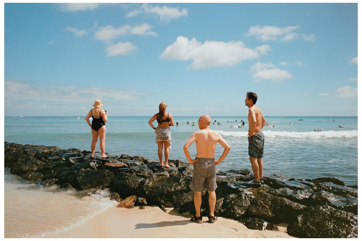 Four people stand on a rocky outcropping at the edge of a beach, looking out at the ocean where many people are swimming and surfing.