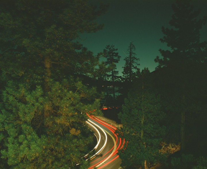 A winding road with light trails from vehicles is visible through pine trees at night.