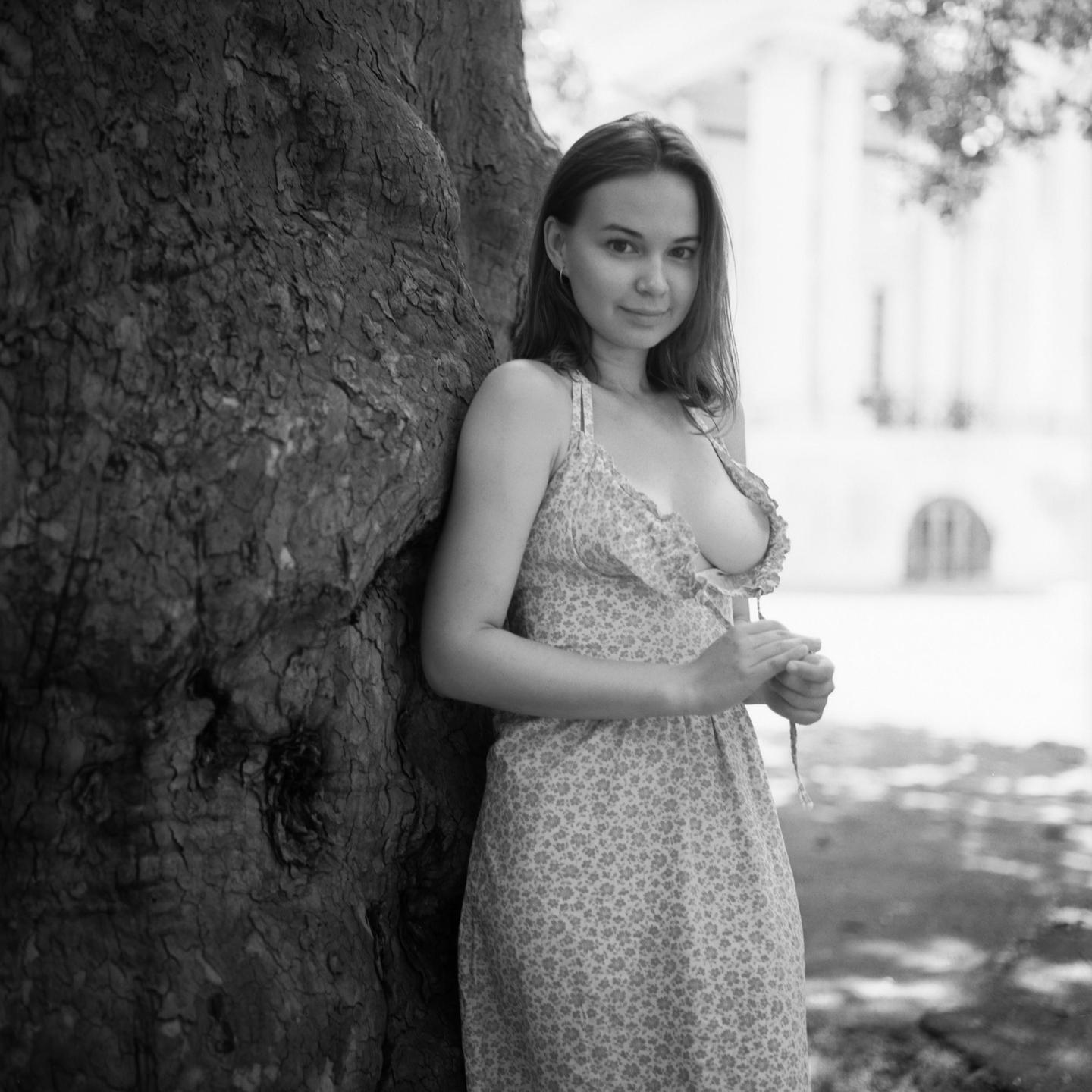 A woman in a floral dress stands next to a tree.