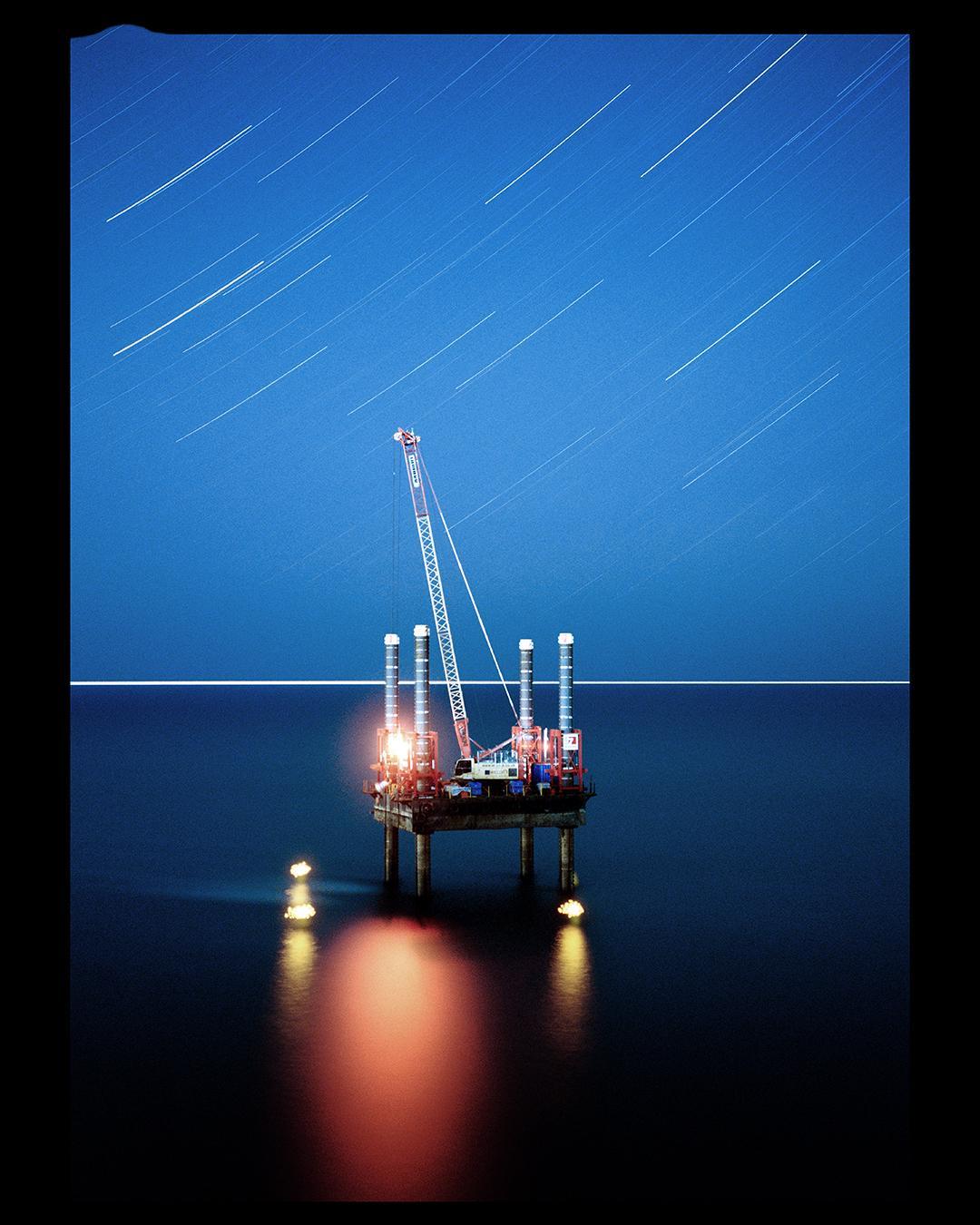 An oil rig with a crane stands in the ocean under a night sky with star trails.