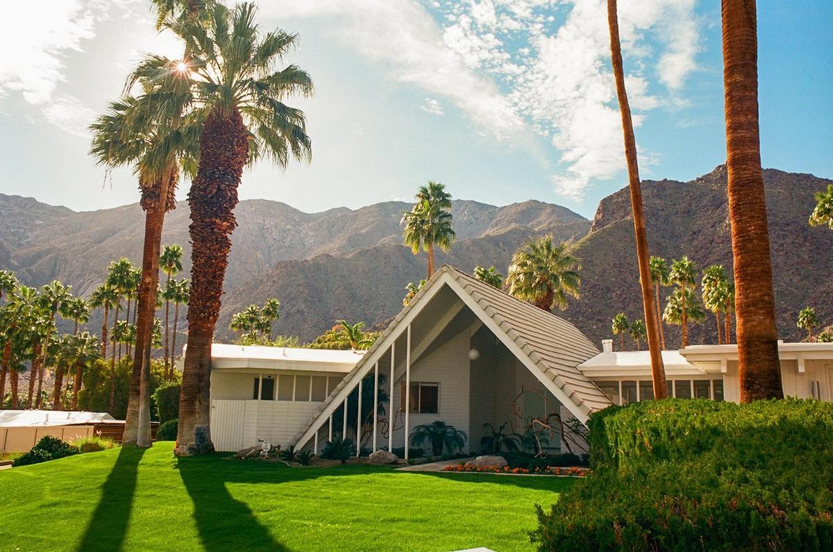 A white A-frame house with a green lawn and palm trees is set against a backdrop of mountains under a bright sky.