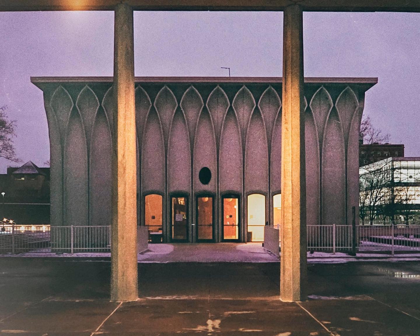A concrete building with arched patterns and illuminated doorways is framed by two large pillars.