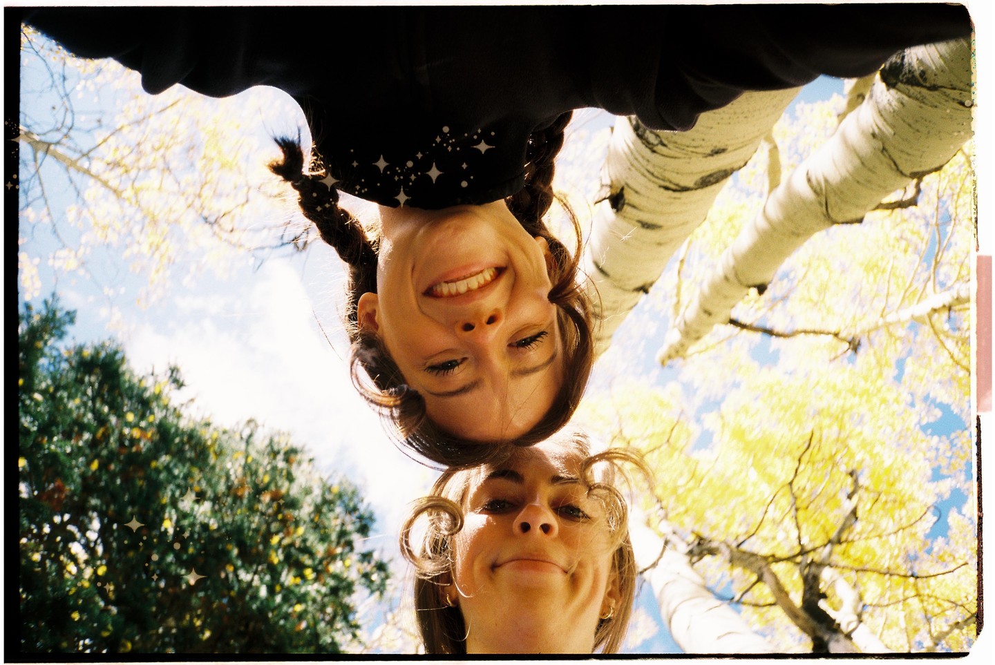 Two women are looking down at the camera, with trees and a blue sky visible in the background.