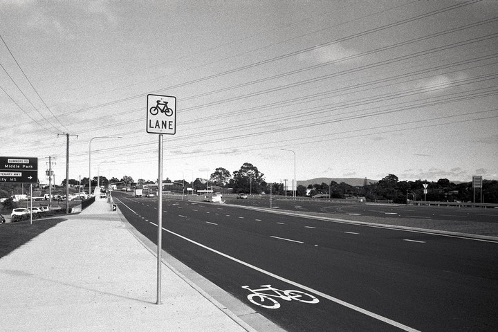 A bicycle lane sign stands beside a road with a painted bicycle symbol on the asphalt, with power lines stretching across the sky.