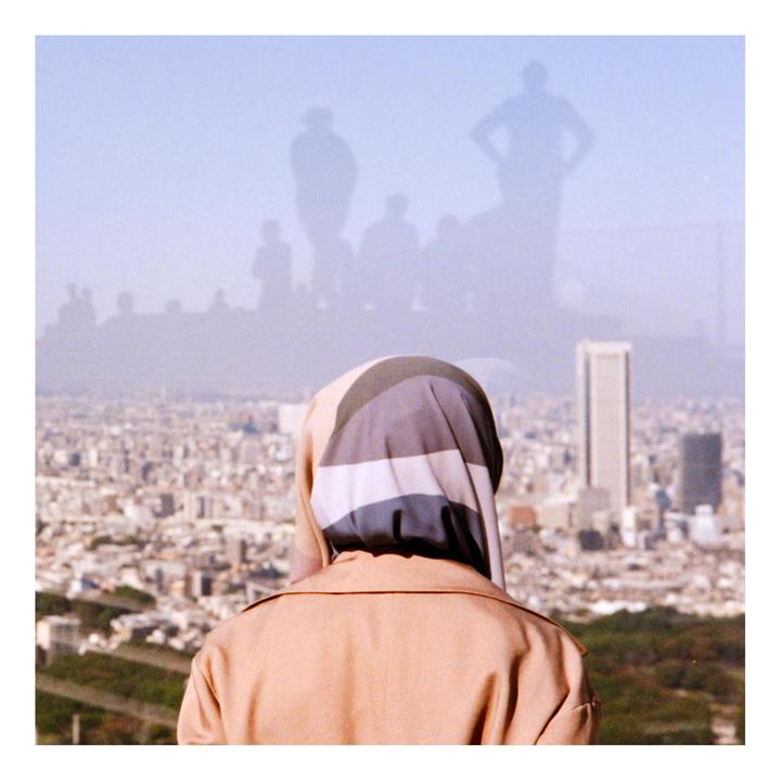 A woman wearing a head covering and a coat looks out over a city with a hazy sky and silhouettes of people.