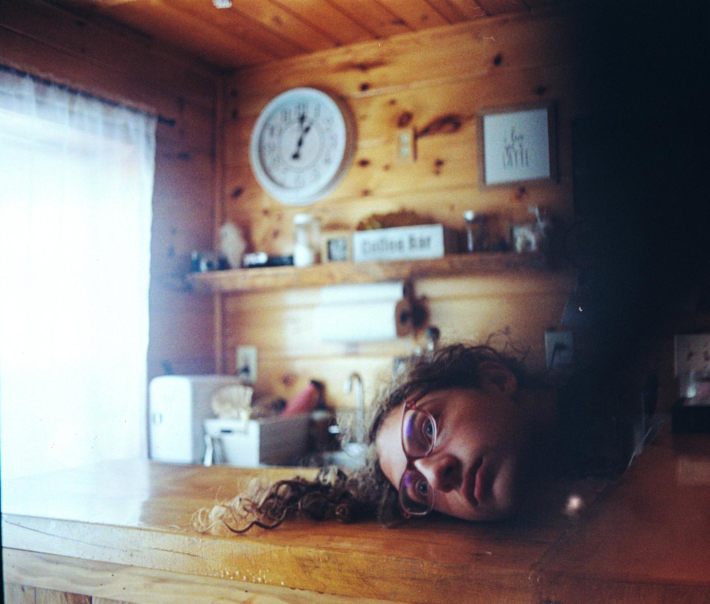 A woman with curly hair and glasses is lying on a wooden counter in a room with wood paneling.