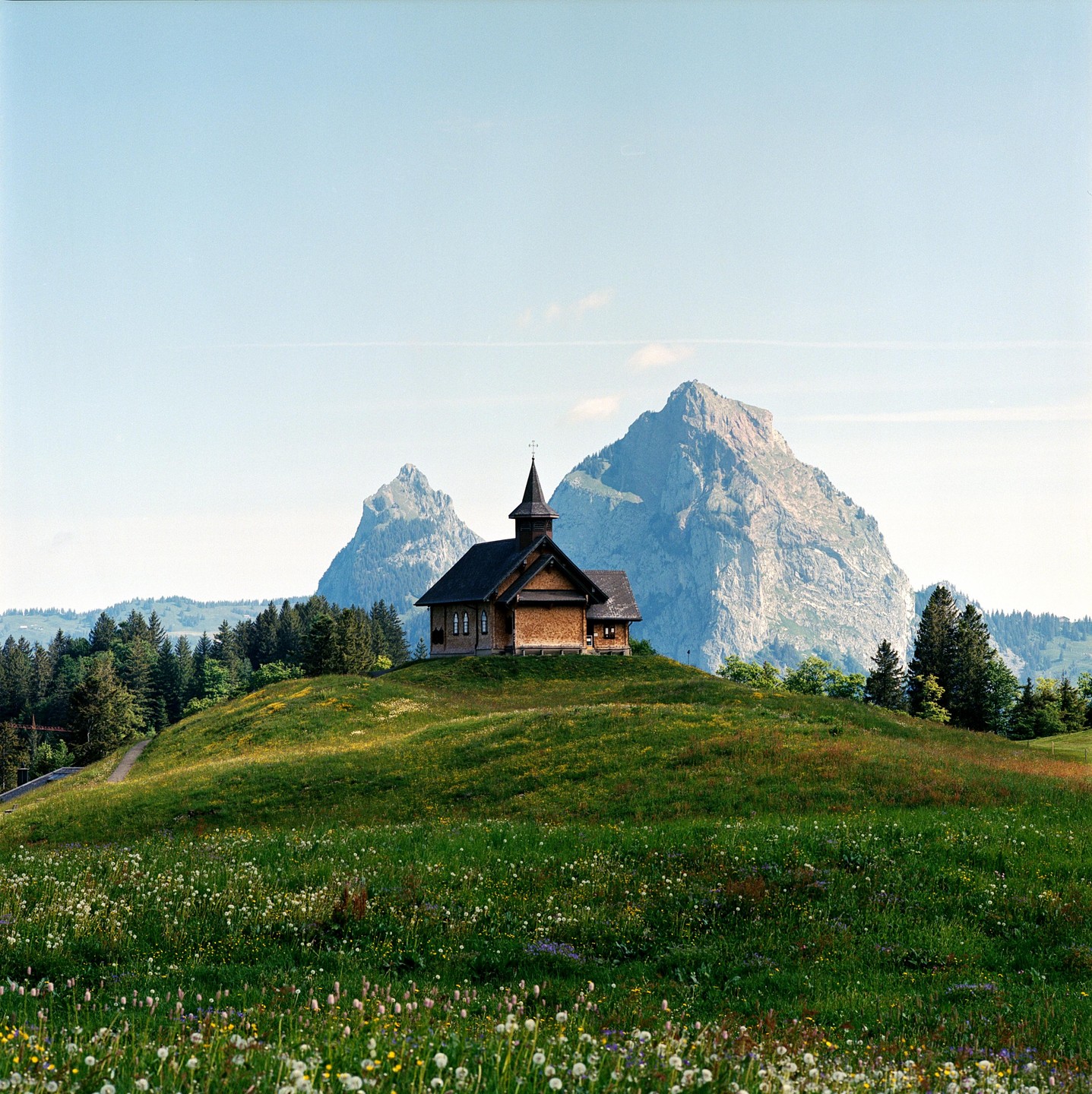 A small wooden chapel stands on a grassy hill with mountains in the background.