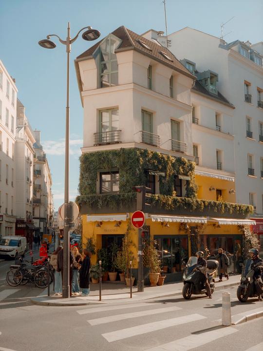 A street corner in Paris features a building with a yellow storefront and a green, leafy facade, with people and motorbikes on the street.