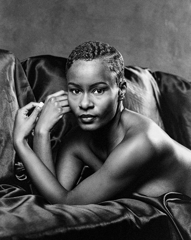 A woman with short, wavy hair is lying on a draped fabric, looking directly at the viewer.