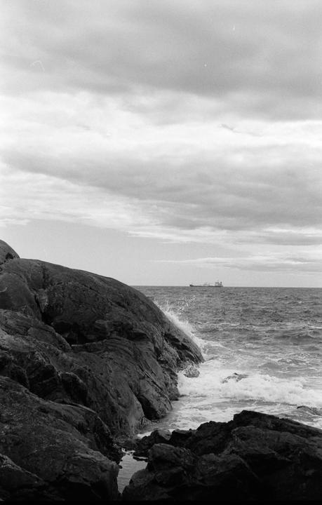 Waves crash against a rocky shore with a ship visible in the distance under a cloudy sky.