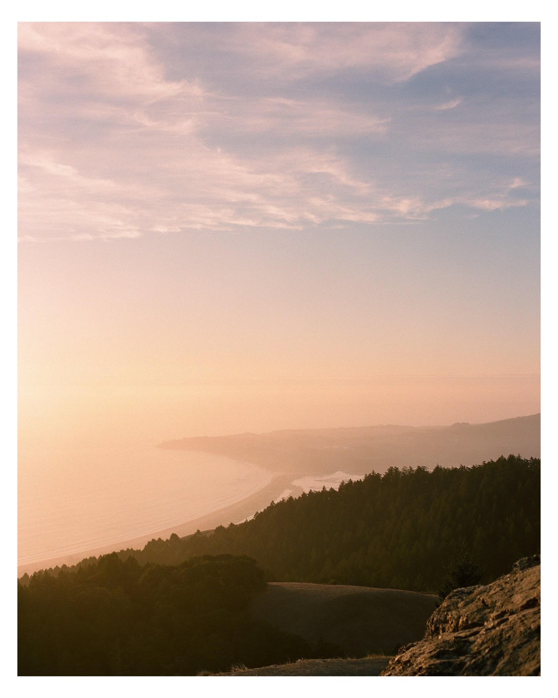 A golden hour landscape shows a forested coastline with a beach and ocean under a hazy sky.