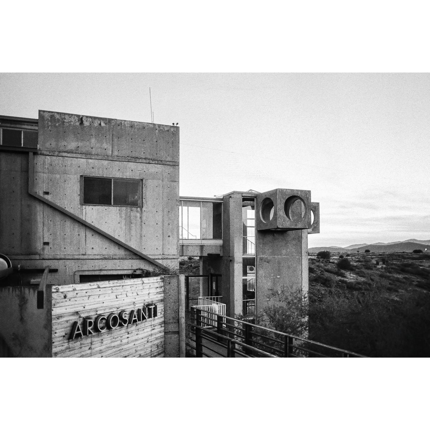 A concrete building with a sign that reads "ARCOSANTI" is situated on a hillside with mountains in the distance.