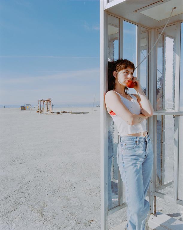 A woman stands in a phone booth on a sandy beach, holding a red telephone receiver to her ear.