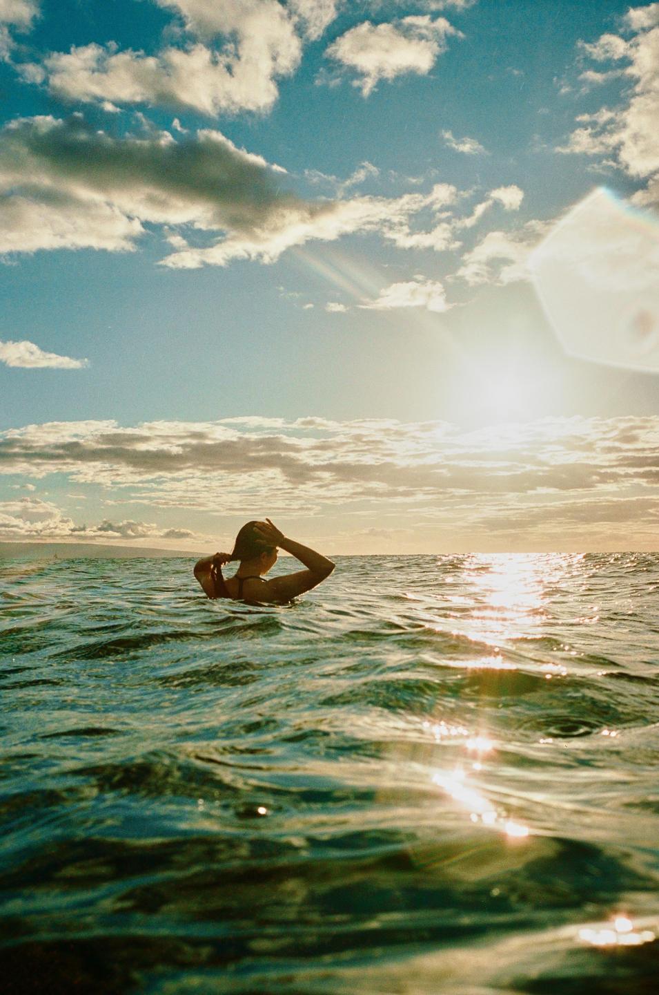 A woman is in the ocean with the sun reflecting on the water and clouds in the sky.