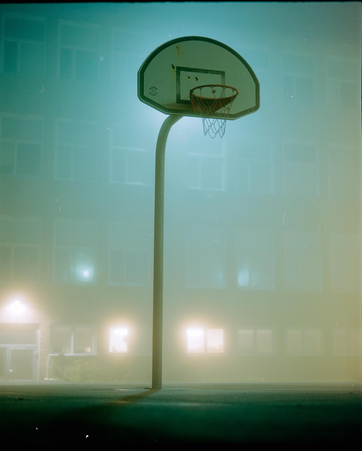 A basketball hoop stands in front of a building with glowing windows on a foggy night.