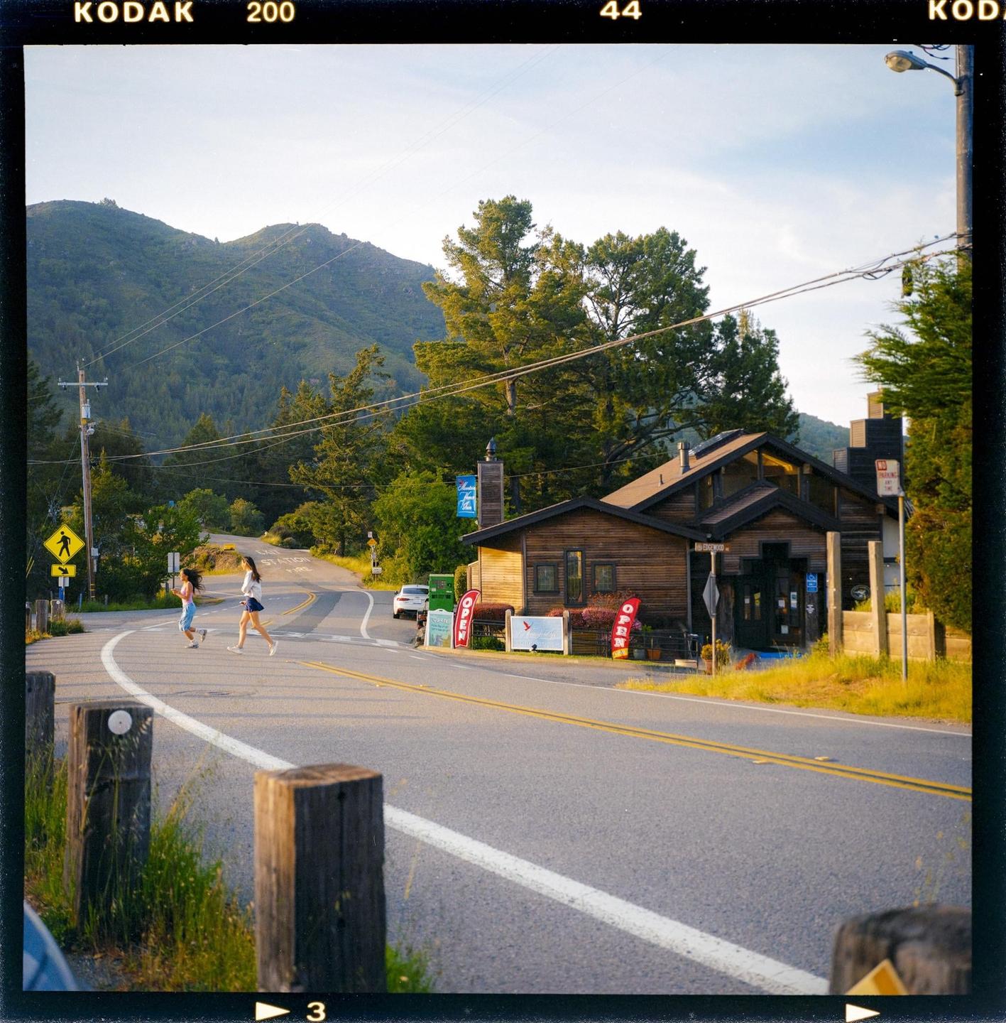 Two women cross a winding road in front of a wooden building with mountains and trees in the background.