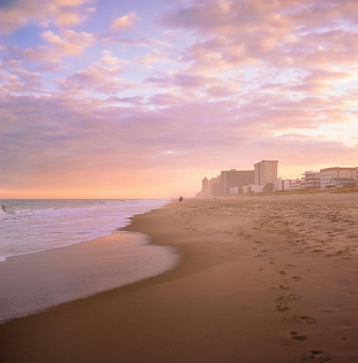 A wide sandy beach with gentle waves and distant buildings is illuminated by the warm glow of sunset.