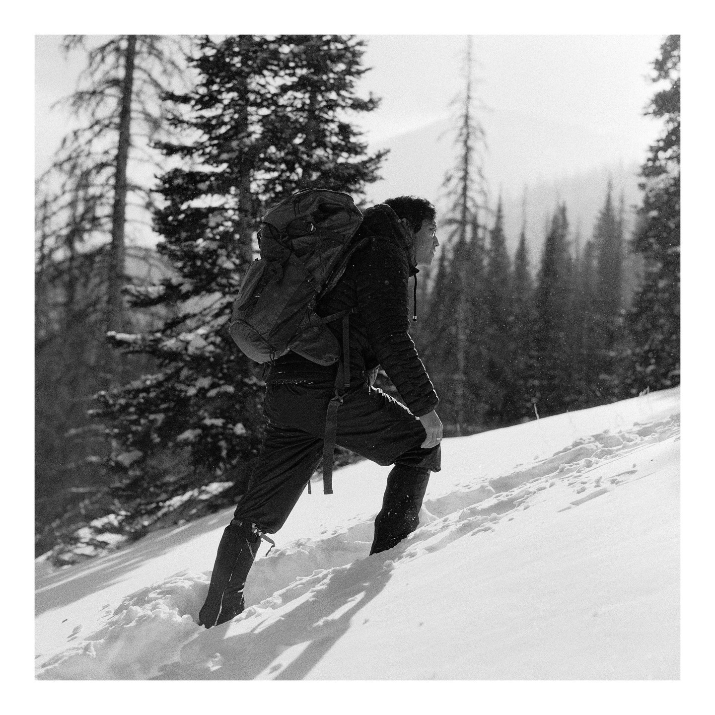 A man with a backpack walks uphill through deep snow in a forest.