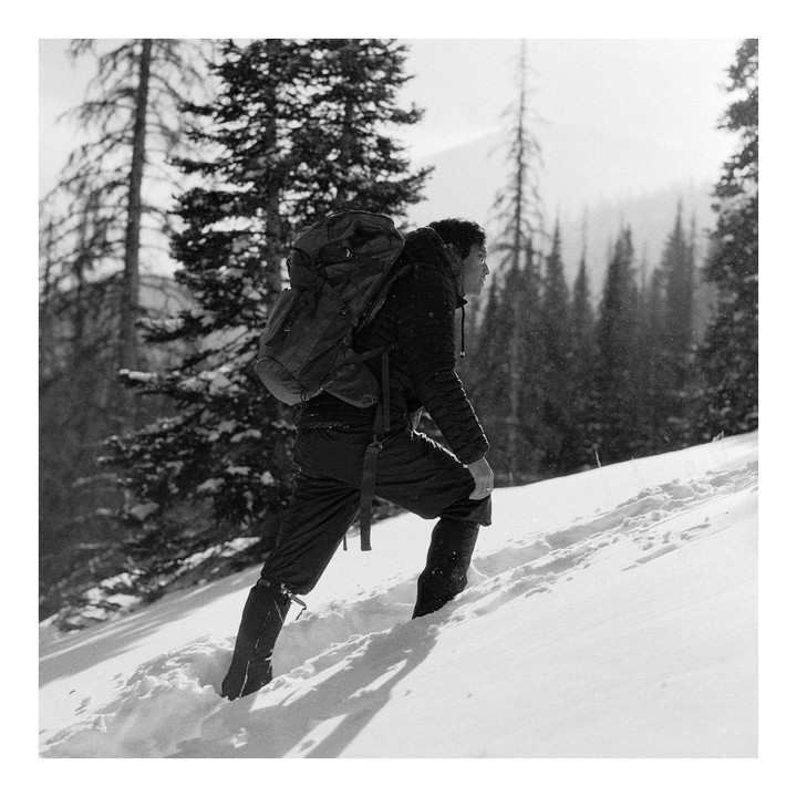 A man with a backpack walks uphill through deep snow in a forest.