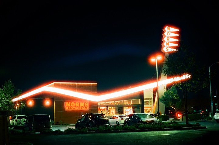 A Norms Restaurant is illuminated by neon lights at night, with cars parked in front.
